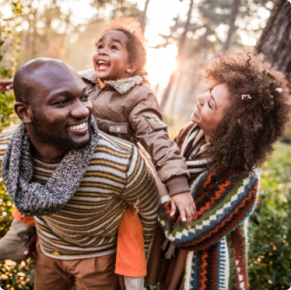 Father carrying young girl on his back as mother looks on during an outdoor walk in the woods.