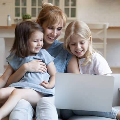 A smiling mother and her two young daughters looking at a laptop computer screen while sitting on the couch.